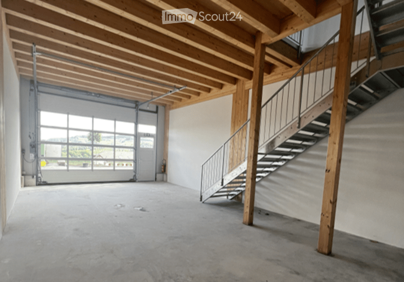 Empty garage with wooden beams, large garage door, and a staircase leading to an upper level.