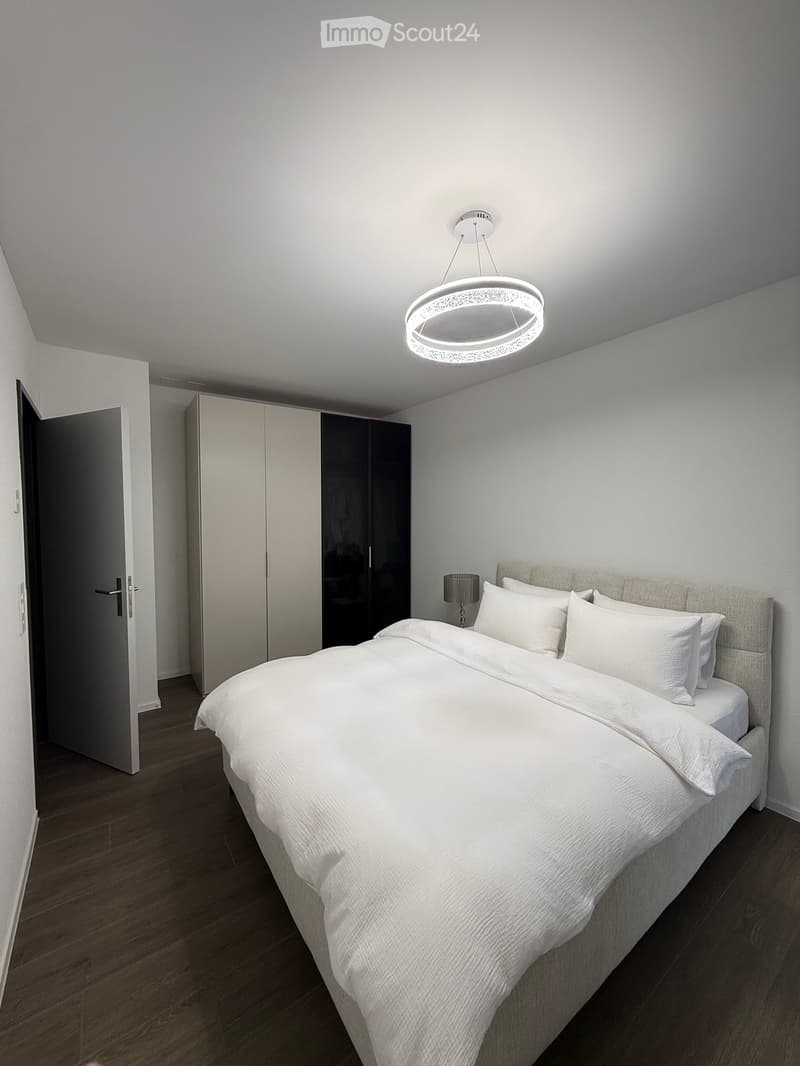 Modern bedroom with a large bed, white bedding, bedside table with lamp, and a sleek wardrobe with mirrored doors.