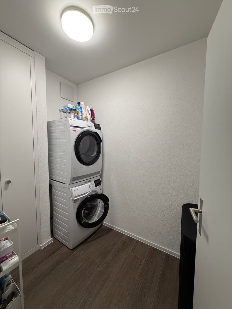 A compact bathroom with a stacked washing machine and dishwasher, wooden flooring, and cleaning supplies on top of the appliances.
