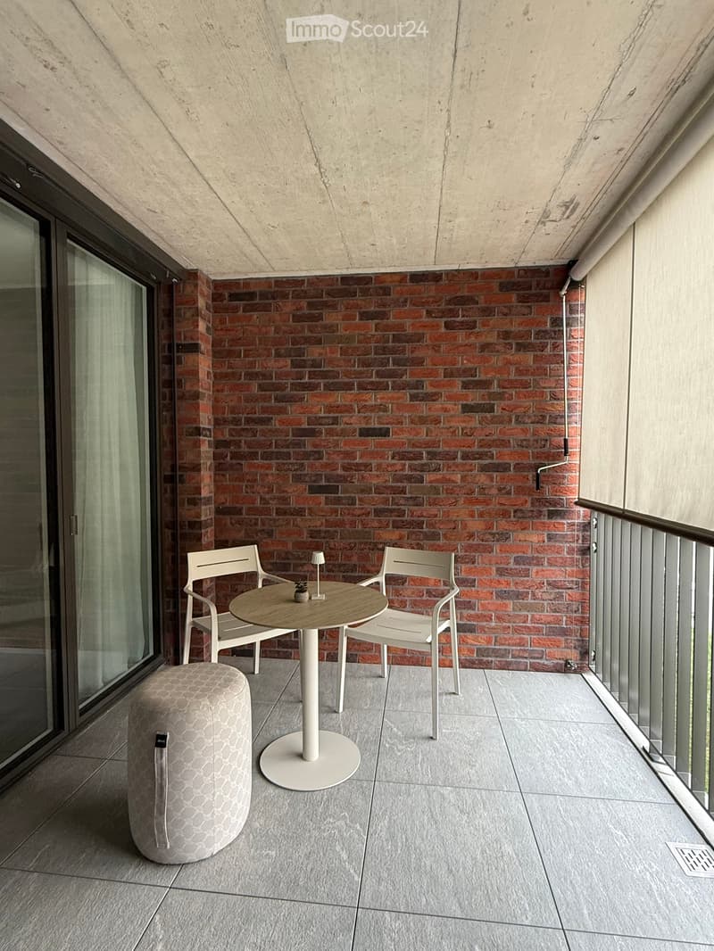 Balcony with a round table, two chairs, and a stool, featuring a brick wall and large glass doors.