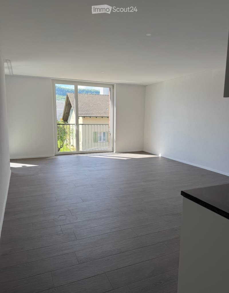 Empty room with wooden flooring, white walls, and a large sliding glass door leading to a balcony with a view of a neighboring building and greenery.