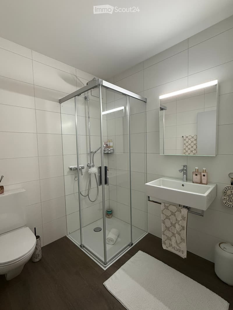 Modern bathroom with white tiles, a glass-enclosed shower, a toilet, a sink with a mirror, and a towel on the floor.