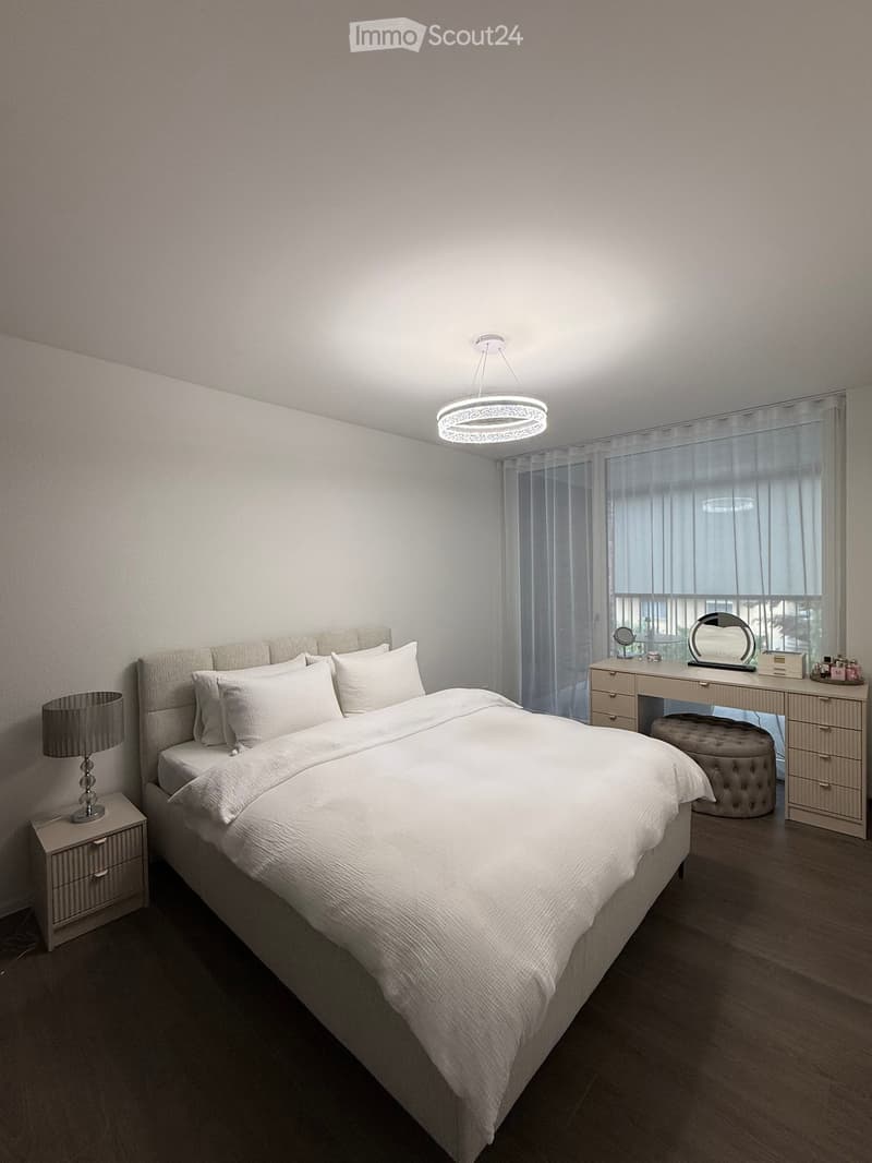 Modern bedroom with a large bed, nightstand with lamp, dresser with mirror, and a round ottoman stool. Large windows with sheer curtains and a modern pendant light fixture.