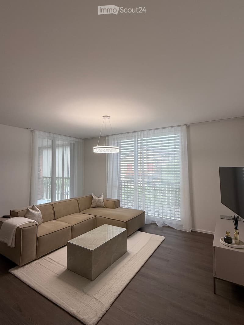 Modern living room with a large sectional sofa, a glass coffee table, a white rug, and a television mounted on the wall. The room features large windows with blinds and a hanging pendant light.