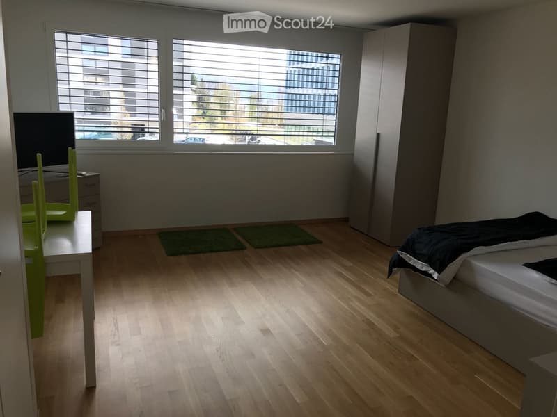 Modern bedroom with wooden flooring, large window with blinds, bed with black bedding, small table with green chair, and wardrobe.
