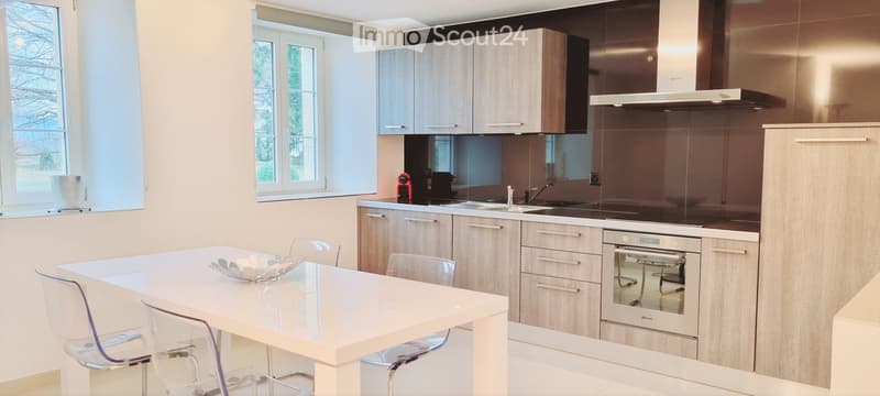Modern kitchen with light wood cabinets, stainless steel appliances, white countertops, and a dining area with a white table and transparent chairs.
