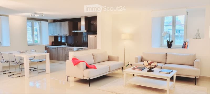 Modern open-plan living and dining area with white walls, light wood flooring, and large windows. Features a white sofa with a red throw, a coffee table with books and a decorative bowl, a dining table with chairs, and a sleek kitchen area with cabinets a
