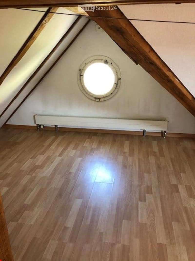Empty attic space with wooden flooring, white walls, exposed wooden beams, and a round skylight window.