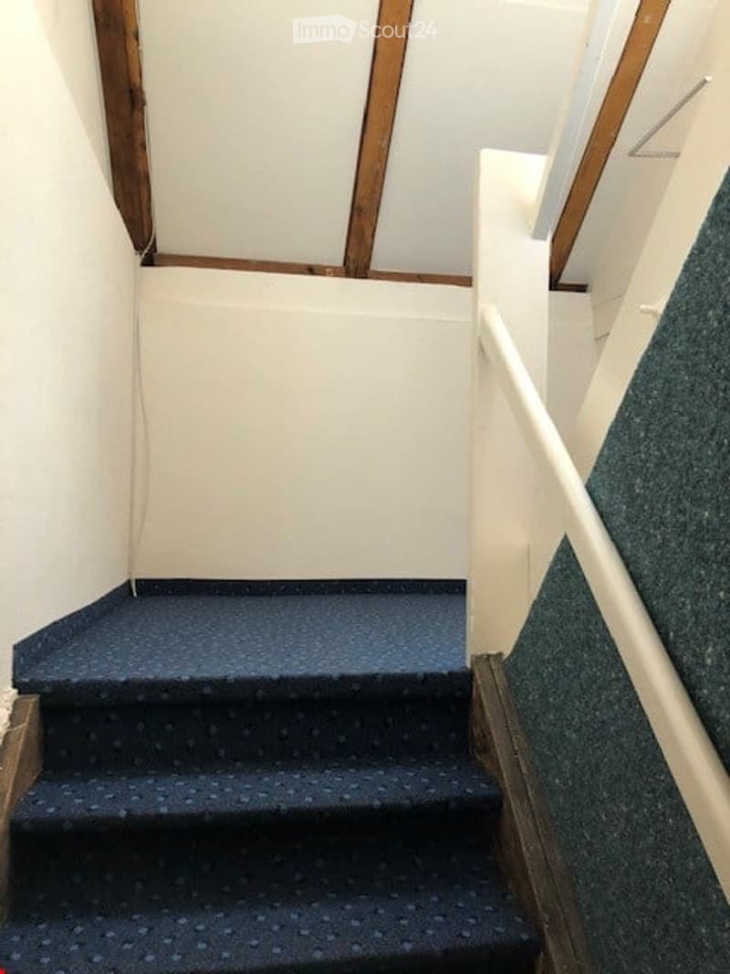 Staircase with blue carpeted steps and white handrail, wooden trim, white walls, and exposed wooden beams on the ceiling.