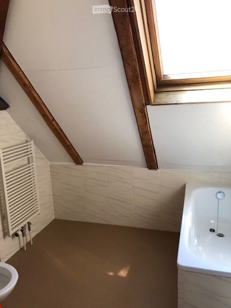 An empty attic bathroom with exposed wooden beams, a white radiator, a bathtub, and a window letting in natural light.