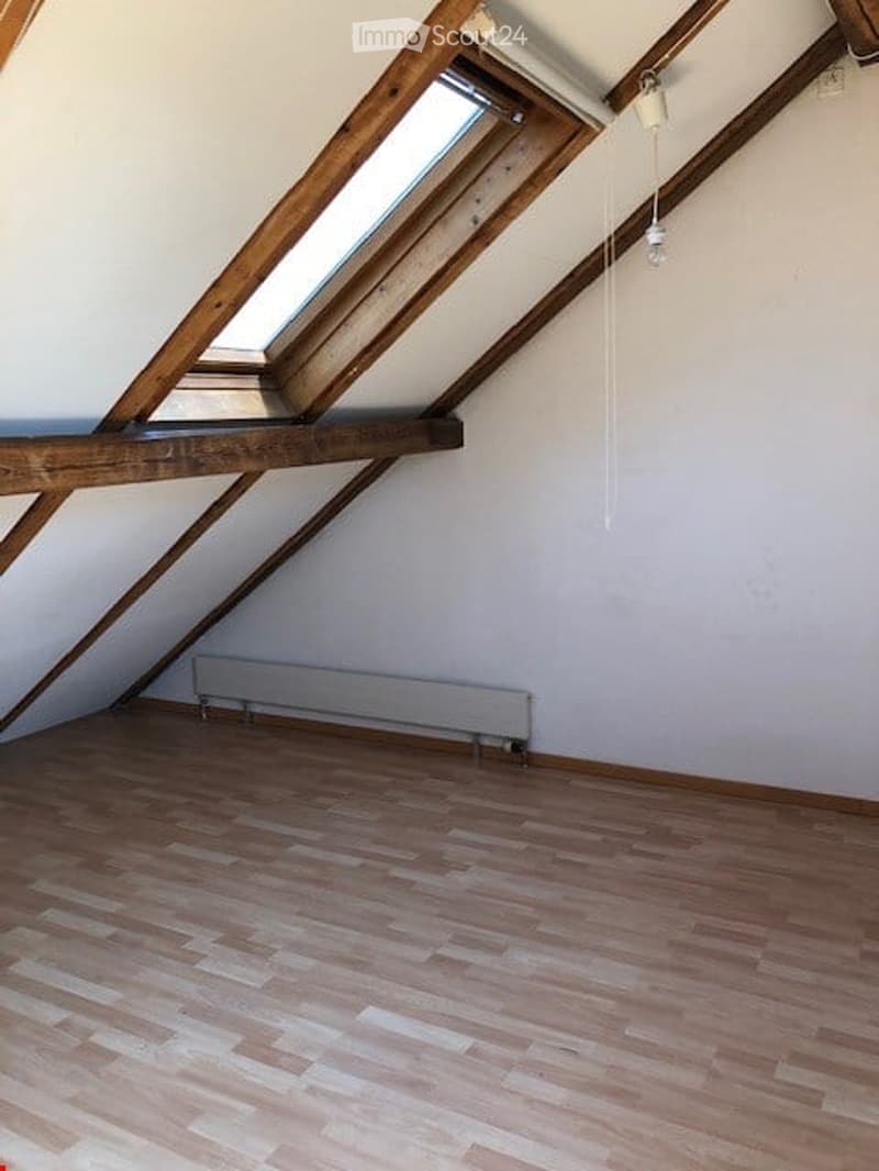 Empty attic room with wooden floor, white walls, exposed wooden beams, skylight windows, and a radiator.