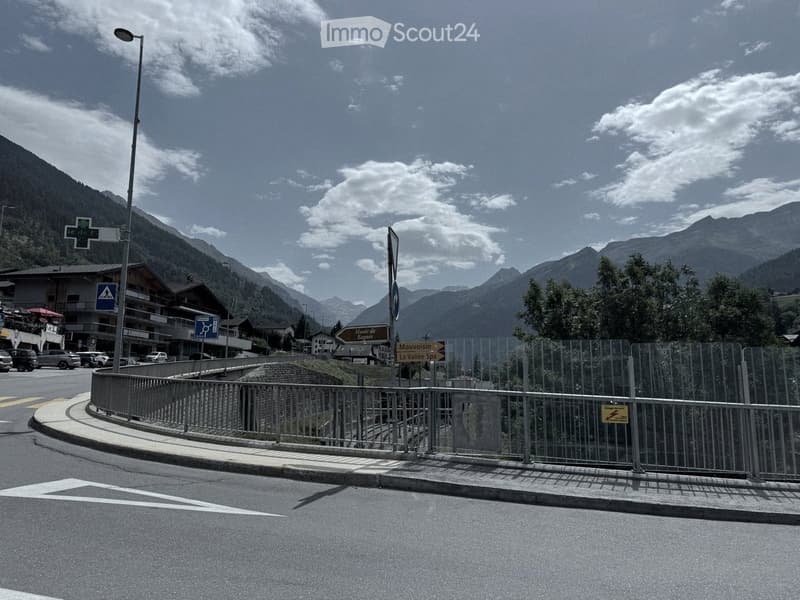 A scenic mountain road with a metal barrier, street signs, and a few parked cars, surrounded by mountains and a partly cloudy sky.