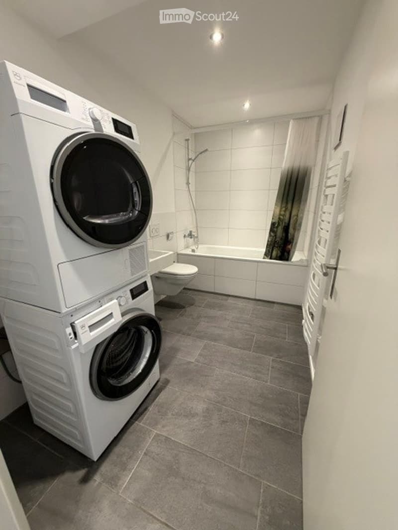 Modern bathroom with grey tiled floor, white tiled walls, a bathtub, toilet, shower, and stacked washing machine and dryer.