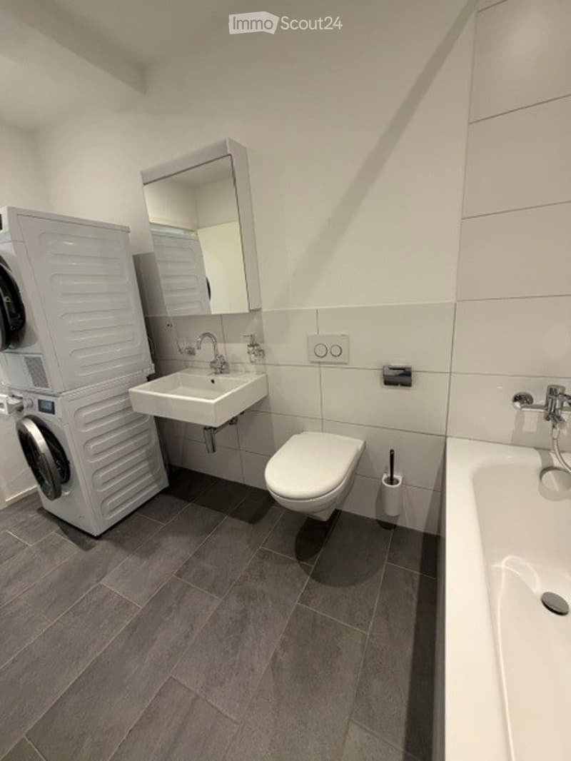 Modern bathroom with a white sink, toilet, bathtub, and a washing machine. Features a large mirror above the sink and a dark grey tiled floor.