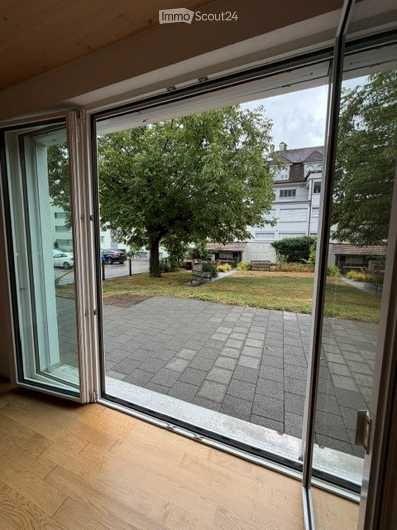 Open sliding glass doors leading to a paved patio area with a view of a green lawn, trees, and neighboring buildings.
