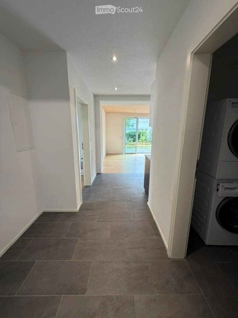 Empty hallway with tiled floor, white walls, and a washing machine visible in an adjacent room.