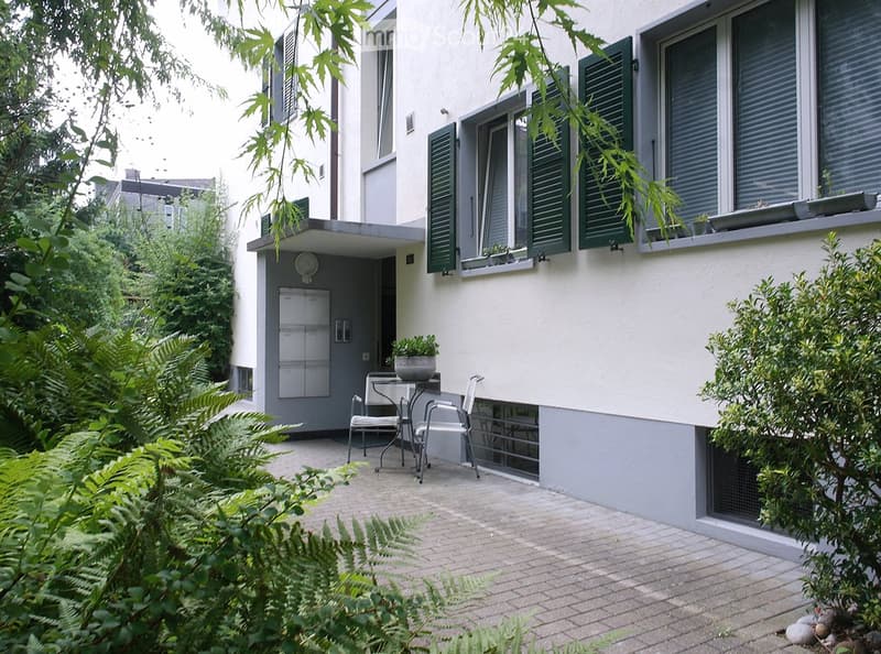 Exterior view with brick floor patio, mailbox, green shutter windows, a table with chairs, and potted plants.