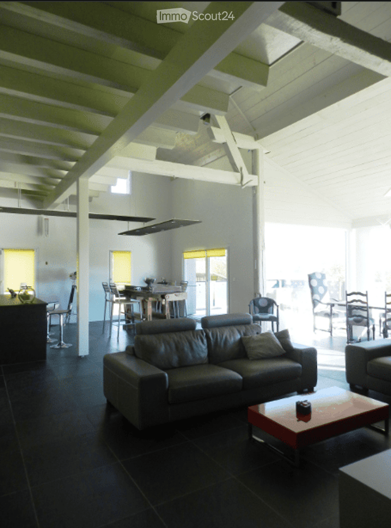 Modern living room with dark tiled flooring, exposed wooden beams on the ceiling, and large windows. Features a dark leather sofa, a red coffee table, and several chairs around a dining table in the adjacent area.