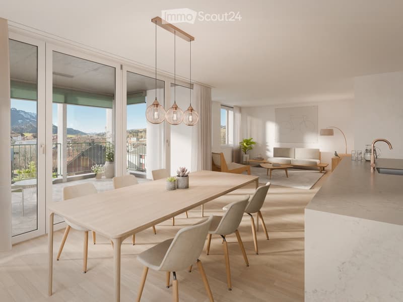 Open-plan living and dining area with a large dining table, modern chairs, and a view of the mountains. The room features a white sofa, a rug, and a coffee table. There are also potted plants and a lamp on the table. The kitchen area is visible with a mar