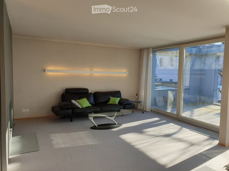 Modern living room with a black leather sofa, glass coffee table, side table, and large windows with curtains. The room features a sleek design with built-in lighting and a view of the balcony.