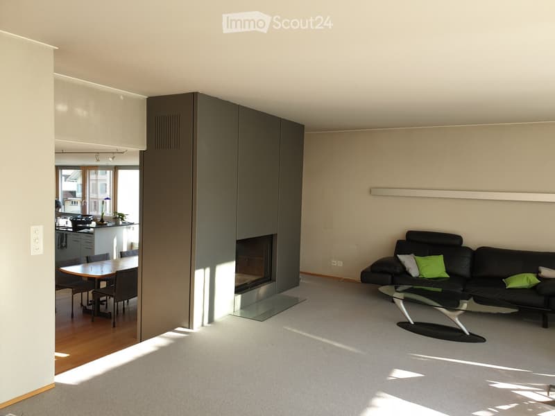 Modern living room with a black leather sofa, glass coffee table, fireplace, dining area with a table and chairs, and a view of the kitchen area.