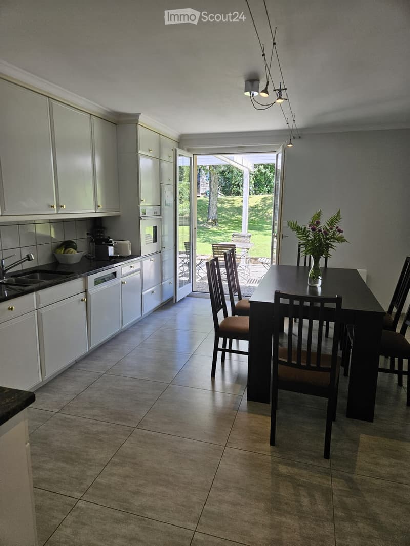 Open-plan kitchen and dining area with white cabinets, a dark dining table with chairs, and a vase of flowers on the table. The kitchen features a sink, oven, and dishwasher, with a view to a garden through large glass doors.