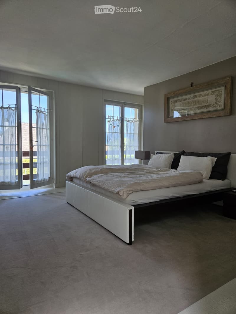 Modern bedroom with a large bed, nightstand, lamp, and picture frame on the wall. Large windows with sheer curtains allow natural light to fill the space.