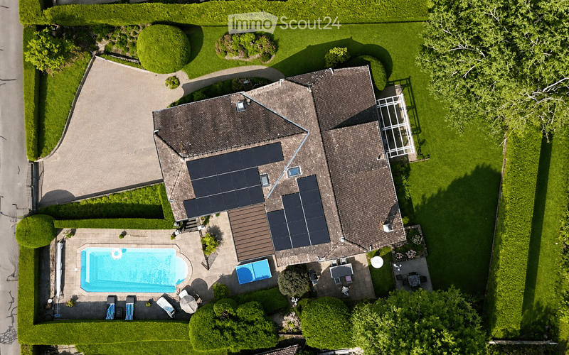 Aerial view of a two-story house with a tiled roof, solar panels, and a well-maintained garden. The property includes a swimming pool, patio furniture, and a driveway.