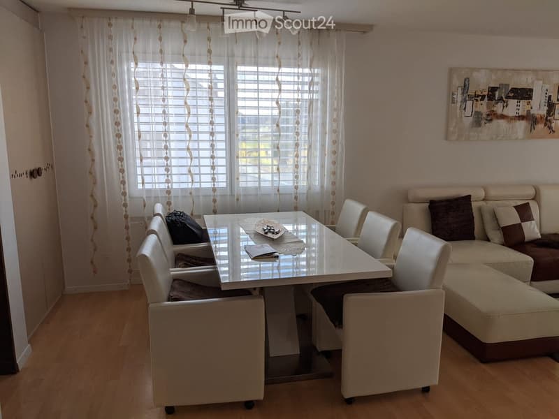 Bright living room with a sofa, coffee table, dining table, chairs, and a handbag on the floor. Large windows with white curtains and a modern painting on the wall.