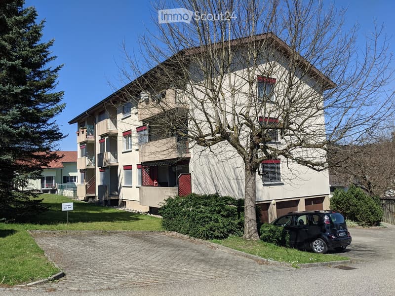 A multi-story residential building with balconies, surrounded by trees and a driveway with a parked car.