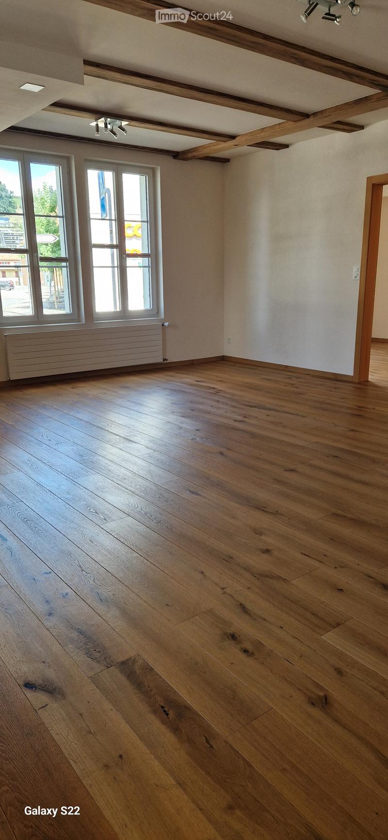 Empty room with wooden flooring, exposed wooden beams on the ceiling, large windows, and a radiator under the windows.