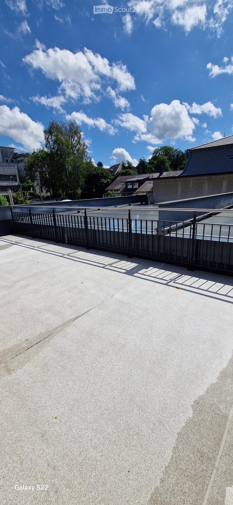 Roof terrace with a concrete floor, metal railing, and a view of surrounding buildings and trees under a partly cloudy sky.