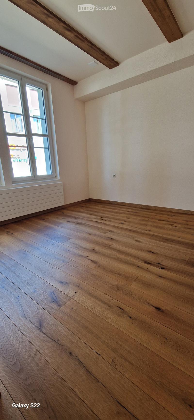 Empty room with wooden flooring, white walls, and a large window with white frame. Exposed wooden beams on the ceiling.