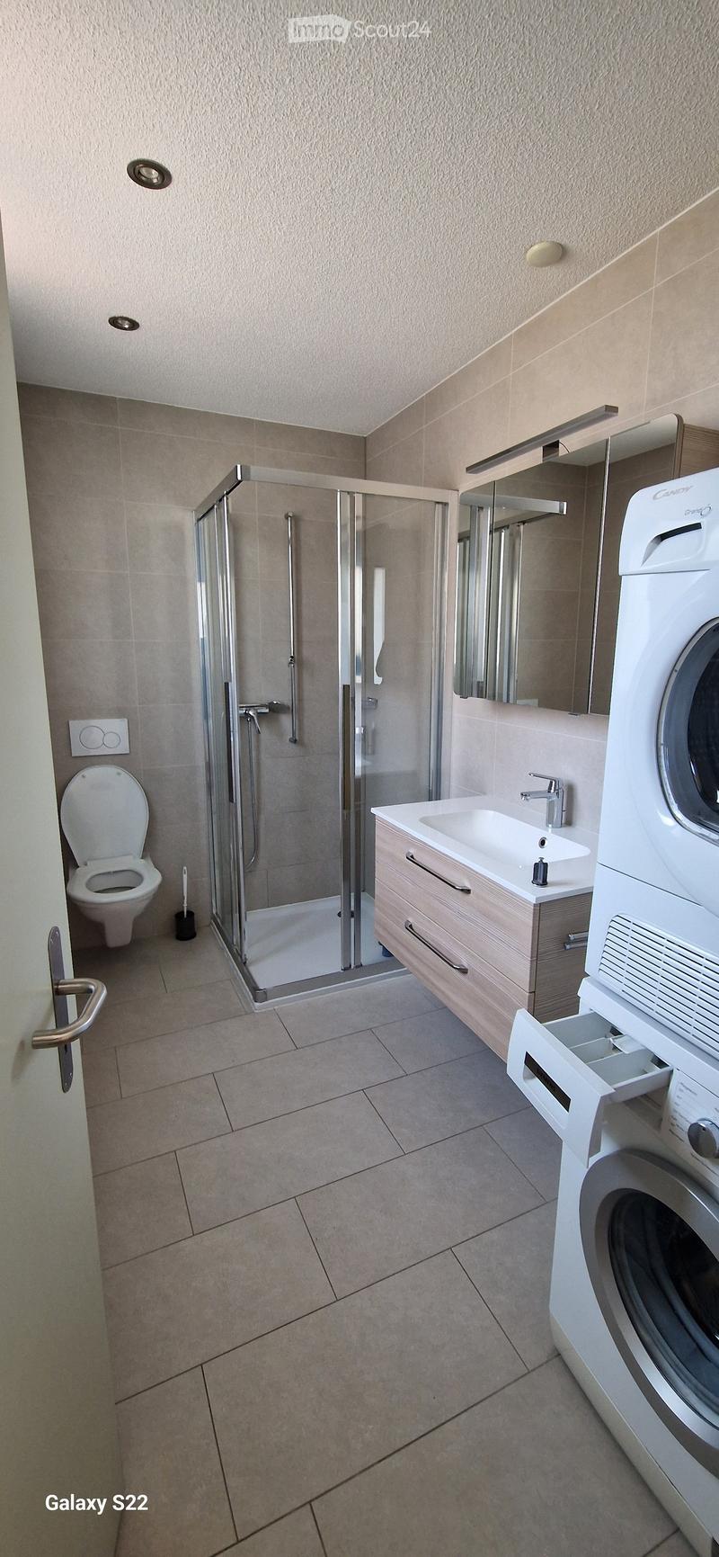 Modern bathroom with a toilet, sink, mirror, glass-enclosed shower, and a washing machine. Light-colored tiles and wooden cabinetry.