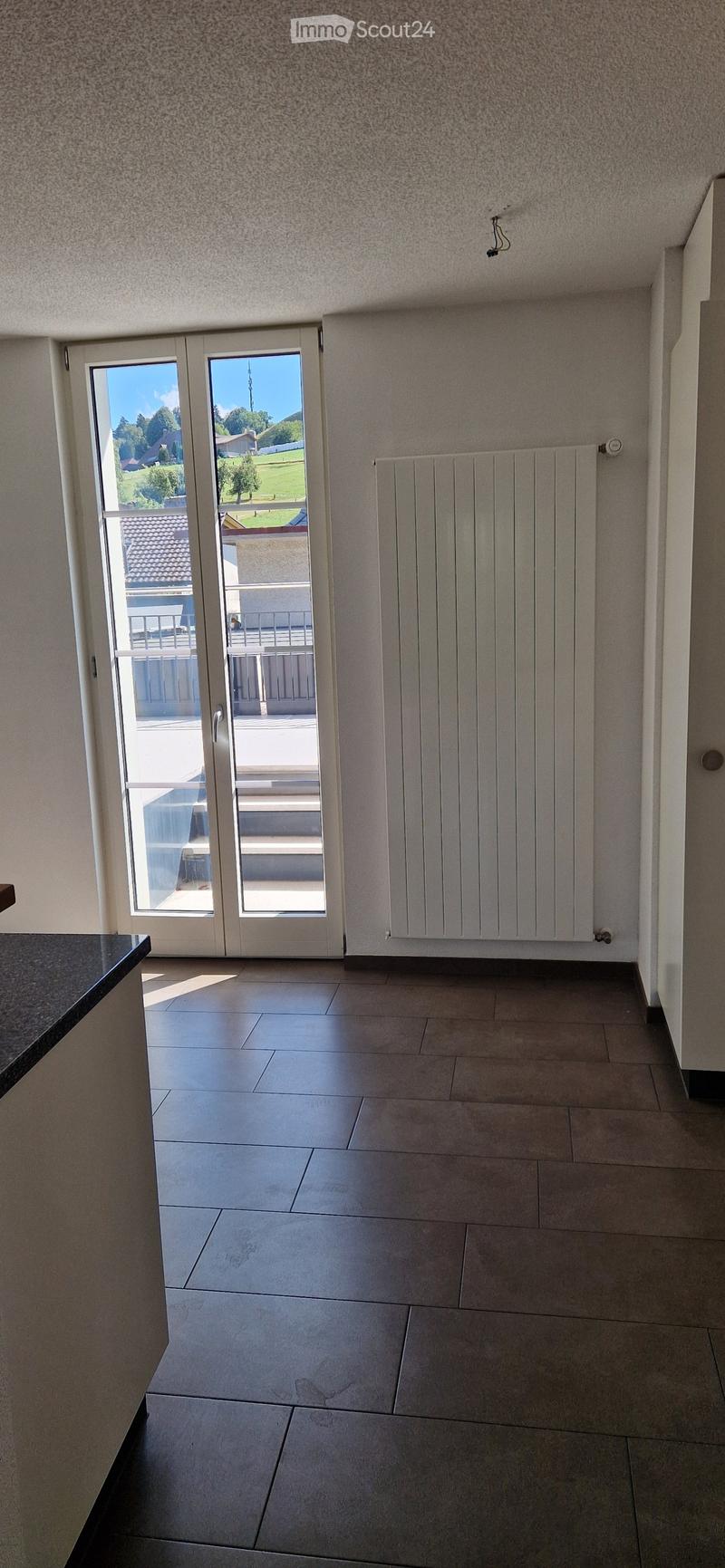 Empty room with tiled floor, white walls, large sliding glass doors leading to a balcony, radiator, and a small black countertop visible on the left side.
