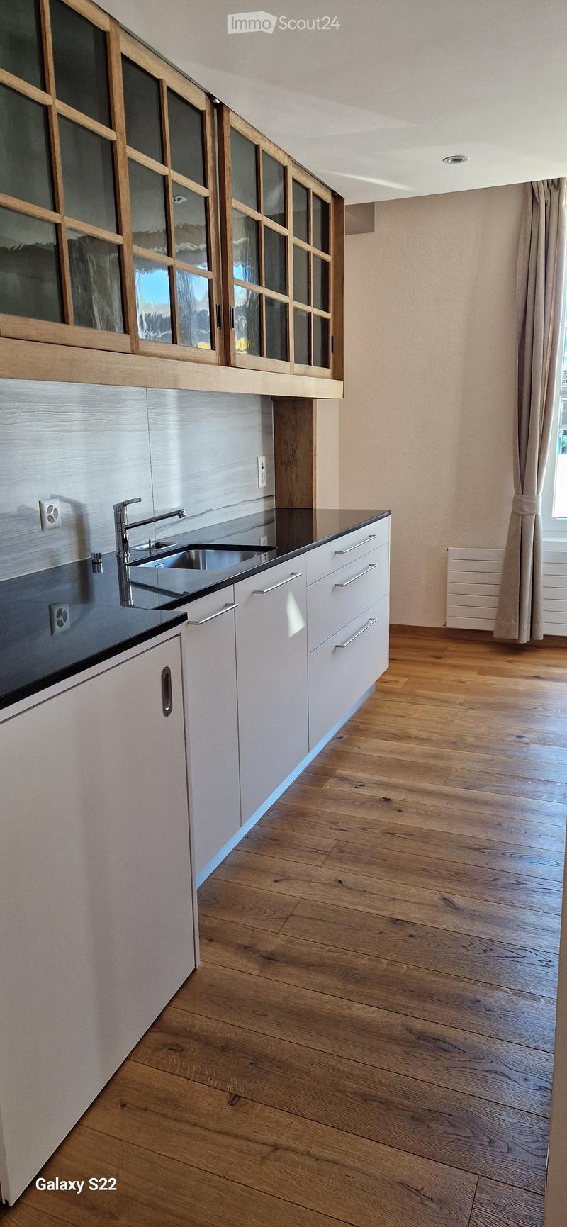 Modern kitchen with white cabinets, wooden flooring, and a black countertop. Features a stainless steel sink and faucet, with large windows and wooden-framed glass panels above the cabinets.