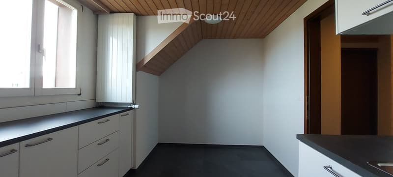 Empty kitchen with white cabinets, dark countertops, and a window letting in natural light. The space features a wooden slanted ceiling and a door leading to another room.