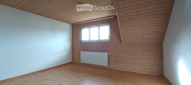Empty room with wooden walls, wooden floor, and a single window with white frame. A radiator is mounted on the wall below the window.