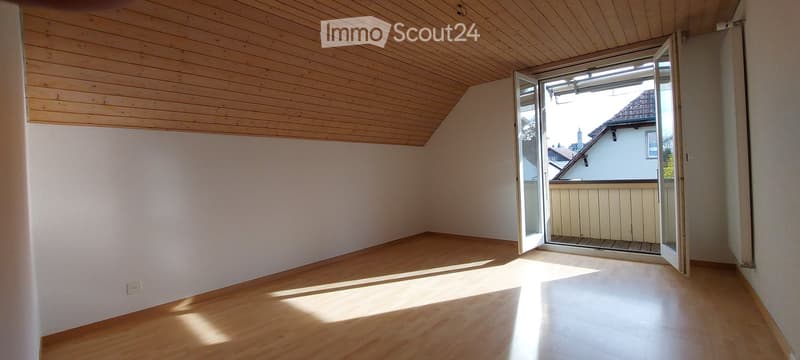 Empty room with wooden flooring and slanted wooden ceiling, featuring an open sliding door leading to a small balcony.