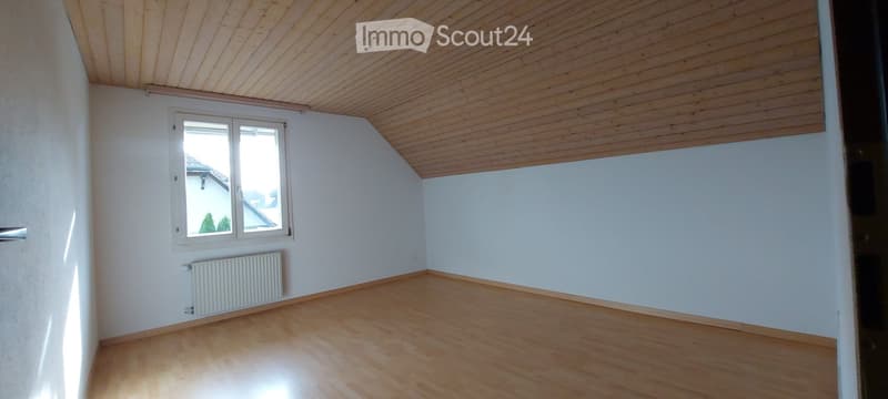 Empty room with wooden floor, white walls, and a slanted wooden ceiling. Features a window with white frame and a radiator below it.