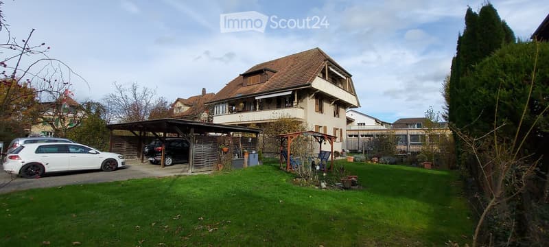 Two-story house with a tiled roof, beige facade, and a covered garage area. The front yard has a well-maintained lawn, a small seating area, and several potted plants. Two cars are parked in the driveway.