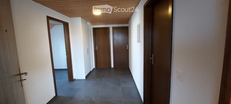 A clean, modern hallway with tiled flooring, white walls, and wooden doors. The ceiling is paneled in light wood, and a single round light fixture provides illumination.