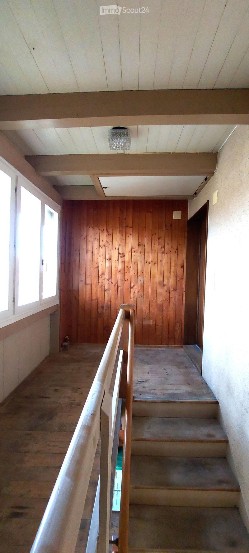 Wooden staircase with a wooden railing, concrete steps, and a wooden door at the end. The walls are painted in a light color, and there is a window on the left side providing natural light.