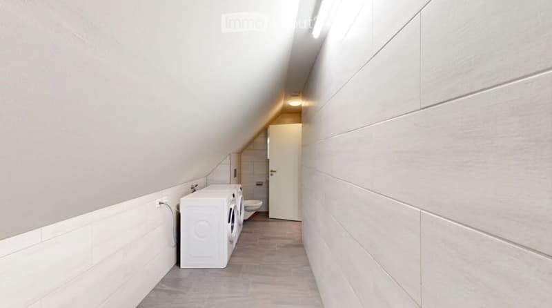 A narrow hallway with a slanted ceiling, white tiled walls, and a wooden floor. A white door is visible at the end, and a small white washing machine is placed against the wall.