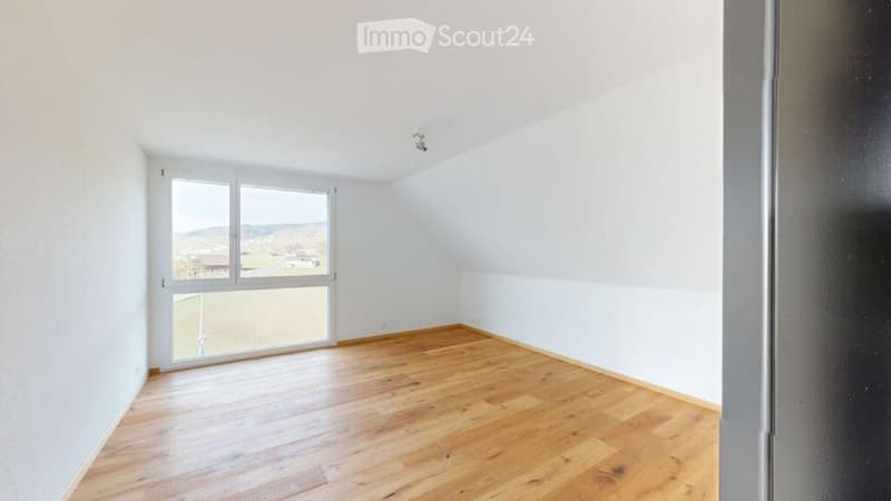 Empty attic room with wooden flooring, large window offering a view, and white walls.