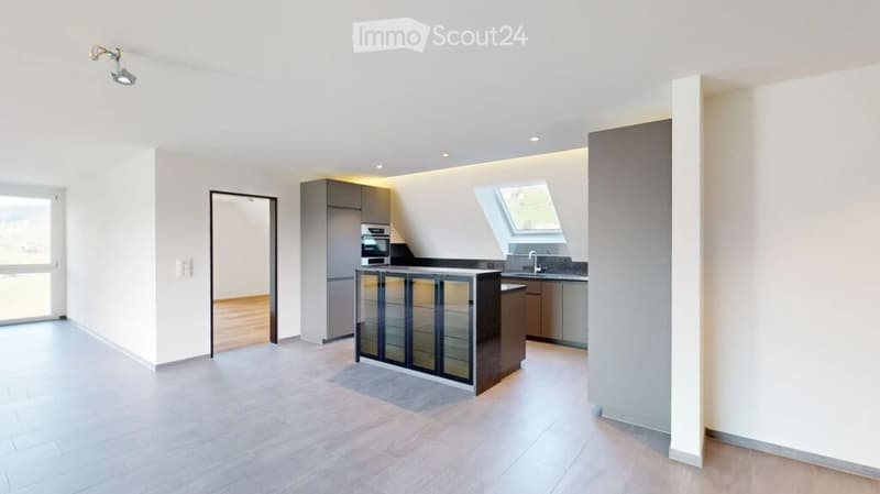 Open-plan kitchen and living area with modern cabinetry, a central island, and large windows. The space is unfurnished, showing only the built-in kitchen units and flooring.