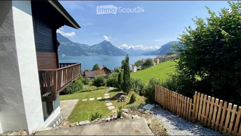 A serene garden area with a wooden fence, stone pathway, and a view of a lake and mountains in the background.