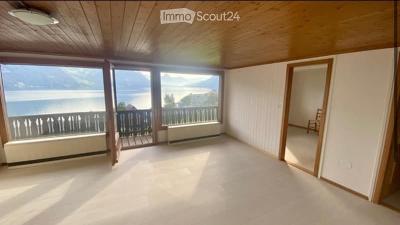 Bright, empty room with wooden ceiling and floor, large windows offering a scenic lake view, and a door leading to a balcony.