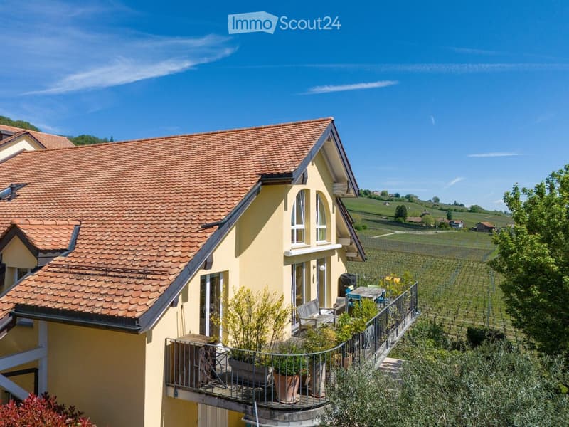 Two-story yellow house with red-tiled roof, large windows, and a balcony with outdoor furniture and potted plants.