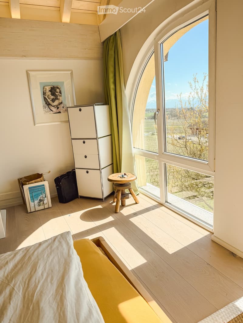 Bright bedroom with wooden flooring, large windows offering a scenic view, a bed with white bedding, a small wooden stool, a white cabinet, and a picture frame on the wall.
